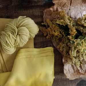 May include: A still-life composition featuring a skein of pale yellow yarn, fabric swatches, and a bouquet of dried yellow flowers. The items are arranged on a dark wooden surface, creating a rustic aesthetic.
