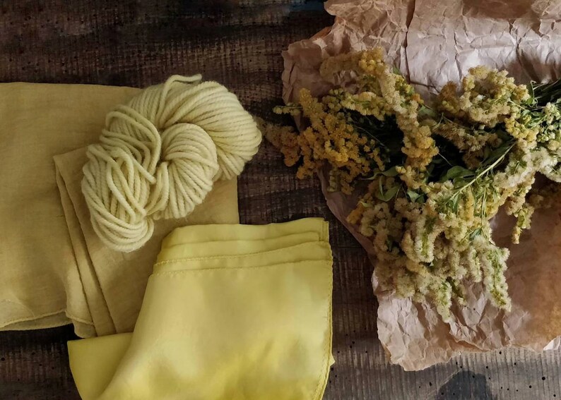May include: A still-life composition featuring a skein of pale yellow yarn, fabric swatches, and a bouquet of dried yellow flowers. The items are arranged on a dark wooden surface, creating a rustic aesthetic.