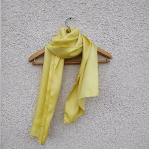 May include: A pale yellow scarf made of a silky material hangs on a wooden hanger. The scarf is draped in a flowing style, with the fabric catching the light and showing subtle variations in color. The background is a textured, off-white wall.