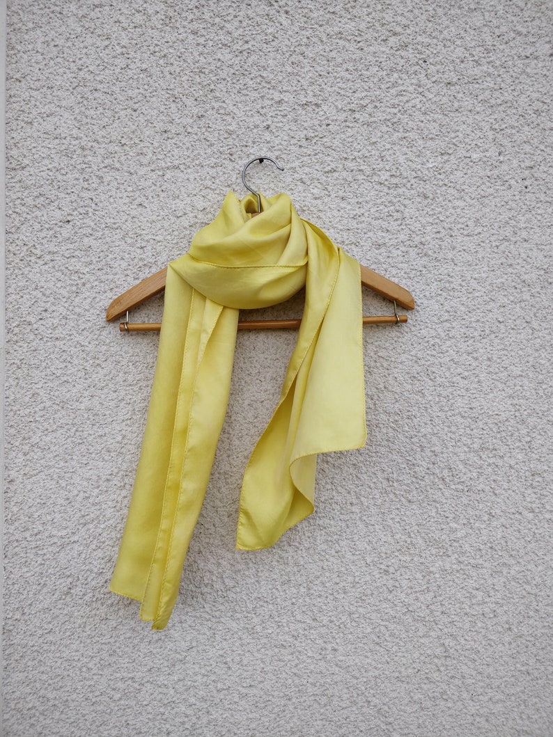 May include: A pale yellow scarf made of a silky material hangs on a wooden hanger. The scarf is draped in a flowing style, with the fabric catching the light and showing subtle variations in color. The background is a textured, off-white wall.