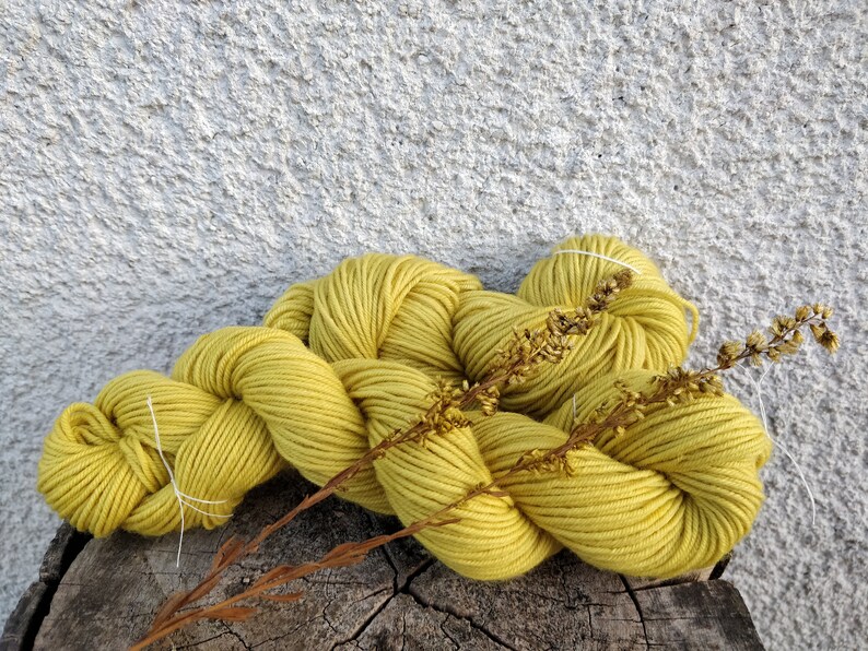 May include: A skein of yellow-green yarn, with a dried plant stem, rests on a weathered wooden surface. The yarn is thick and twisted, with a soft, textured appearance. The background is a textured, light gray wall.