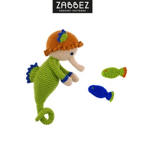May include: A green crocheted seahorse with an orange hat and blue trim. The seahorse is looking at two small crocheted fish, one green and one blue.