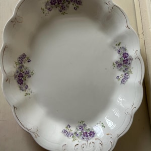May include: A white ceramic serving bowl with a scalloped edge and gold trim. The bowl features three floral clusters of purple flowers with green leaves arranged symmetrically around the interior rim and embossed detailing.