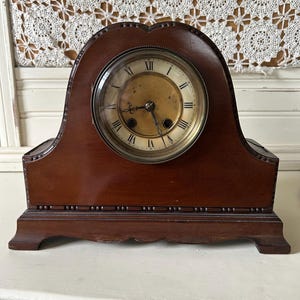 May include: A dark brown wooden mantel clock with an arched top and a circular face. The clock features a gold-toned dial with black Roman numerals, two dark metal hands, and two winding holes positioned at the four and eight o'clock marks.