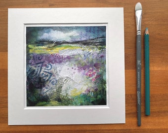 May include: A framed abstract watercolour painting with a landscape scene. The painting features a cloudy sky, a field of purple flowers, and a green hillside in the background. The painting is framed in a white mount.