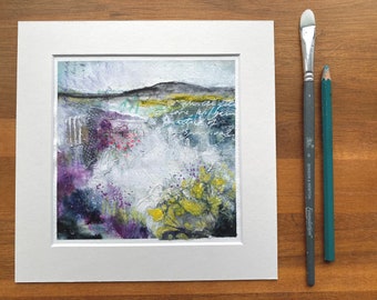 May include: A framed abstract painting with a white background and a landscape scene in shades of purple, green, yellow, and blue. The painting includes the text "in the world get a little bit of perspective and survival".