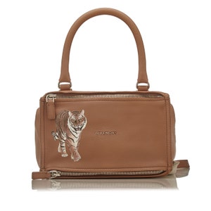 May include: A brown leather handbag with a top handle and a tiger embroidered on the front. The bag has a gold zipper and the word "Givenchy" is printed on the front.