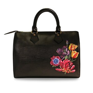 May include: Black leather handbag with a floral design featuring pink, yellow, and blue flowers. The bag has a top handle and a zipper closure.