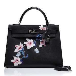 May include: Black leather handbag with a floral print featuring pink flowers and butterflies. The bag has a silver lock and a top handle.
