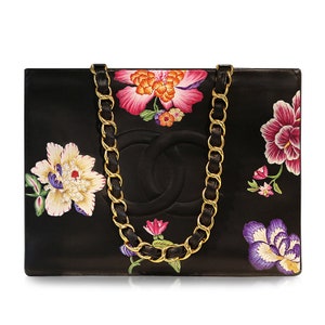 May include: A black leather tote bag with a gold chain strap and floral embroidery. The bag features a large Chanel logo and a variety of colorful flowers.