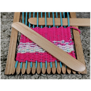 May include: A wooden loom with a pink and blue woven tapestry. The loom has a wooden frame and a set of wooden dowels. A wooden weaving tool is laying across the loom.