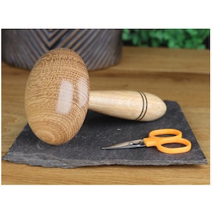 Peut inclure: Un outil en bois en forme de champignon avec une poignée, utilisé pour presser les coutures dans les projets de couture. L'outil est sur une surface en ardoise noire avec une paire de ciseaux à manches orange.