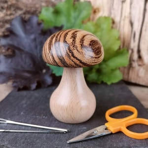 May include: A wooden mushroom-shaped needle minder with a dark brown and light brown striped top and a light brown base. Two needles and a pair of orange scissors are on a black slate surface.
