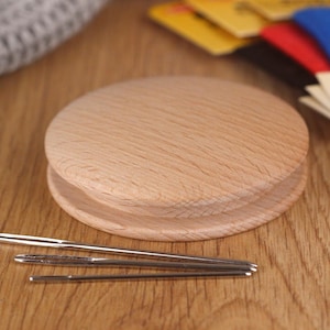 May include: A wooden needle minder with a round shape and a smooth, light brown finish. Three needles are placed on top of the minder.
