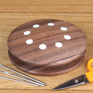 May include: A wooden spool with eight white dots around the edge. The spool is on a wooden surface with three needles and a pair of orange scissors.