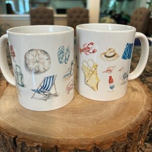 May include: Two white ceramic mugs with beach-themed illustrations. One mug features a sand dollar, beach chair, and flip-flops. The other mug displays a lobster, swimsuit, and hat. Both mugs have a curved handle and are set on a wooden surface.