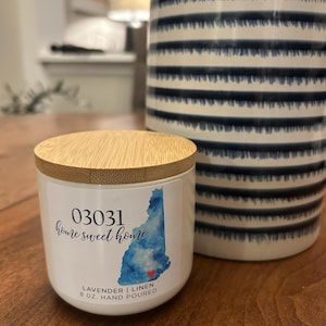 May include: A white candle with a wooden lid, featuring the text "03031 home sweet home" and a blue watercolor map of New Hampshire. The candle is labeled "Lavender | Linen 227g. Hand Poured". A blue and white striped ceramic jar is in the background.