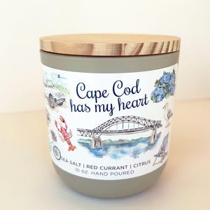 May include: A matte taupe candle in a cylindrical container with a wooden lid. The label features the text 'Cape Cod has my heart' and 'SEA SALT | RED CURRANT | CITRUS 10 OZ. HAND POURED' alongside watercolor illustrations of a bridge, lobster, and car.