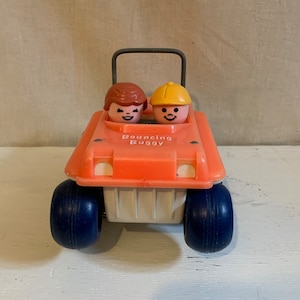 Puede incluir: Un coche de juguete naranja vintage con dos figuras sonrientes en el interior. El coche tiene ruedas azules grandes y el texto "Bouncing Buggy" en la parte delantera. El juguete es de plástico y tiene un asa.
