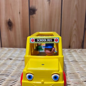 Könnte beinhalten: Ein gelber Spielzeug-Schulbus aus Plastik mit einem lächelnden Gesicht, blauen Augen und roten Rädern. Der Bus hat ein Schild mit der Aufschrift "SCHOOL BUS" und eine kleine Figur eines Menschen auf dem Fahrersitz.
