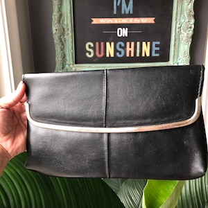 Puede incluir: Clutch negra con marco de metal plateado. El bolso rectangular tiene un cierre de solapa y está dividido en tres secciones por costuras verticales. El fondo incluye un letrero enmarcado que dice "I'M WALKING ON SUNSHINE."