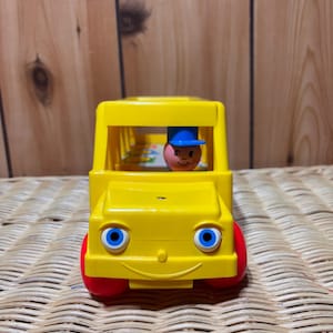 May include: A yellow toy school bus with a smiling face, blue eyes, and red wheels. A small figure with a blue hat is sitting in the driver's seat.