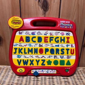 May include: A red and yellow VTech Little Smart Phonics toy with a handle. The toy has a large yellow letter board with the alphabet and pictures of objects that start with each letter. The toy also has a button that plays sounds and a volume control knob.