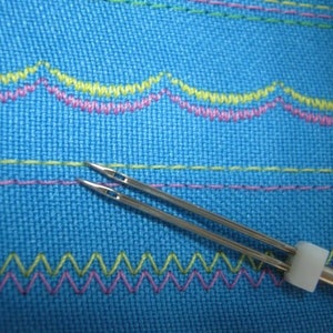 Puede incluir: Primer plano de una herramienta de aguja de doble punta con un mango de pl&aacute;stico blanco. La herramienta de aguja est&aacute; sobre una tela azul con costuras de colores.