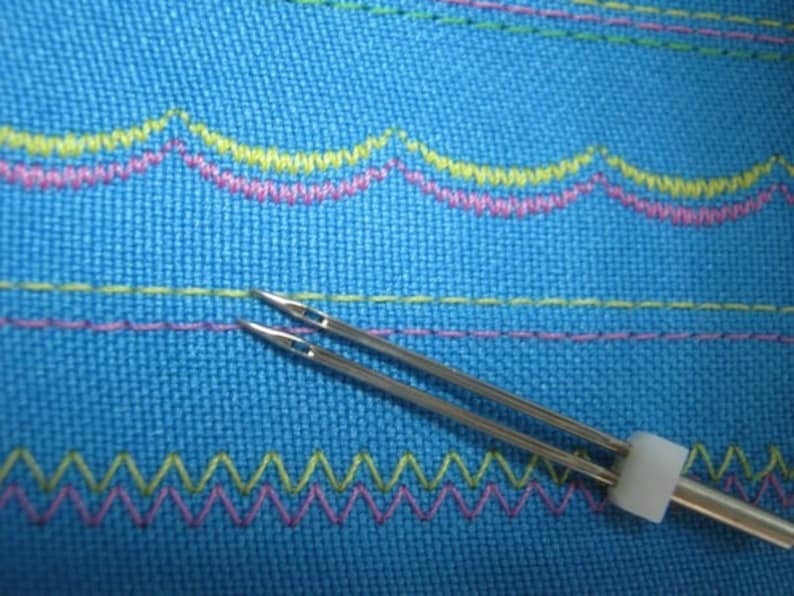 Puede incluir: Primer plano de una herramienta de aguja de doble punta con un mango de pl&aacute;stico blanco. La herramienta de aguja est&aacute; sobre una tela azul con costuras de colores.