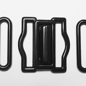May include: Black metal bra strap slider with two loops on each side. The slider has a rectangular shape with a small, rectangular opening in the center.