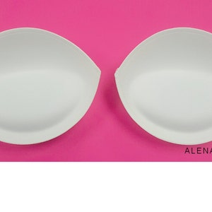 Puede incluir: Dos insertos de sujetador blancos, con forma de l&aacute;grima, sobre un fondo rosa.