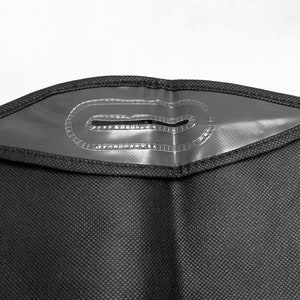 Pu&ograve; includere: Primo piano di un porta abiti nero con un'apertura per la maniglia rinforzata. L'apertura della maniglia &egrave; un rettangolo di plastica trasparente con un bordo cucito. Il materiale della borsa ha un motivo sottile e strutturato.