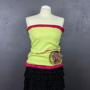 Puede incluir: Un top verde lima sin tirantes con un ribete de volantes rosa y un adorno floral. El top se combina con una falda negra de volantes en capas.