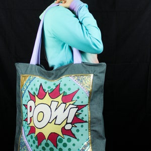 May include: A gray canvas tote bag with a colorful comic book style graphic that says "POW!" in a yellow and white font. The graphic is surrounded by a gold trim and a purple and blue glitter trim.