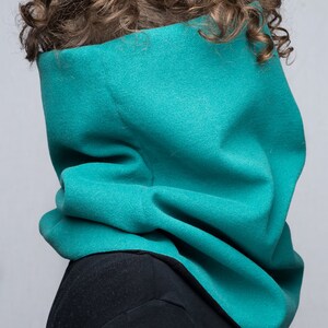 May include: A teal blue fleece neck warmer with a wide, folded collar. The neck warmer is worn by a person with curly brown hair.