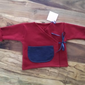 Può includere: Un maglione rosso per neonato con una tasca blu e un nastro blu. Il maglione ha uno stile avvolgente.