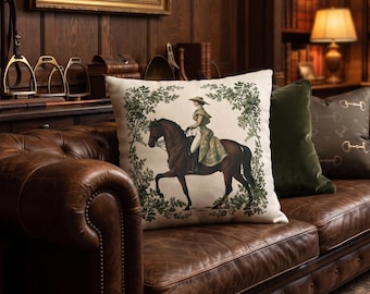 May include: A square throw pillow featuring a central illustration of a person riding a brown horse, framed by green foliage. The pillow is placed on a brown leather tufted sofa alongside a solid dark green pillow and a dark brown patterned pillow.