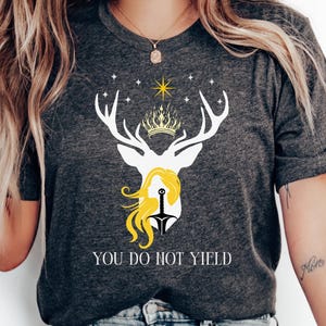 Puede incluir: Una camiseta gris con una silueta de ciervo blanco, una corona dorada y una mujer de pelo amarillo sosteniendo una espada. El texto "You do not yield" está impreso debajo de la imagen.