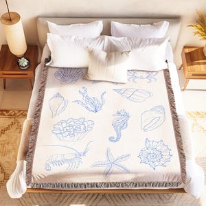 May include: A beige throw blanket with blue seashell and sea creature illustrations, including a seahorse and starfish, covers a bed with white pillows. The blanket has a fringed edge and is placed on a wooden bed frame.