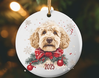 May include: A round, white Christmas ornament featuring a golden doodle dog with a red bow and festive greenery. The year 2025 is printed in red. The ornament hangs from a gold string against a blurred background of Christmas lights and greenery.