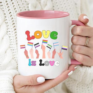 Puede incluir: Taza de cerámica blanca con interior y asa rosa. La taza presenta las palabras "Love is Love" en letras coloridas, junto con ilustraciones de manos sosteniendo varias banderas del orgullo. La taza es sostenida por una persona.