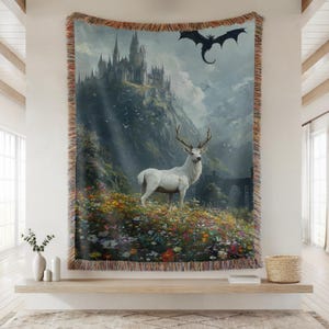 May include: A woven tapestry featuring a fantasy landscape with a white stag in a field of flowers, a castle on a mountain, and a dragon in the sky. The tapestry has a fringed border and is displayed on a wall.