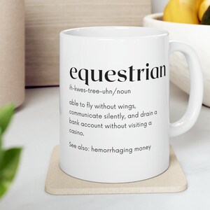 以下が含まれることがあります： 白いセラミック製のマグカップに、黒い文字で「equestrian」という言葉の定義が印刷されています。定義は次のとおりです。「ih-kwes-tree-uhn/名詞 翼なしで飛ぶことができ、静かにコミュニケーションを取り、カジノに行かずに銀行口座を空にすることができる。参照: 資金の流出」