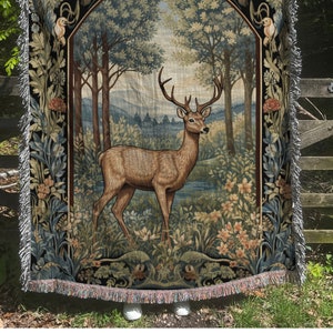 May include: A tapestry throw blanket with a vintage design featuring a deer standing in a forest. The deer is brown with large antlers and is surrounded by green trees and flowers. The tapestry is framed by a black border with intricate floral designs.