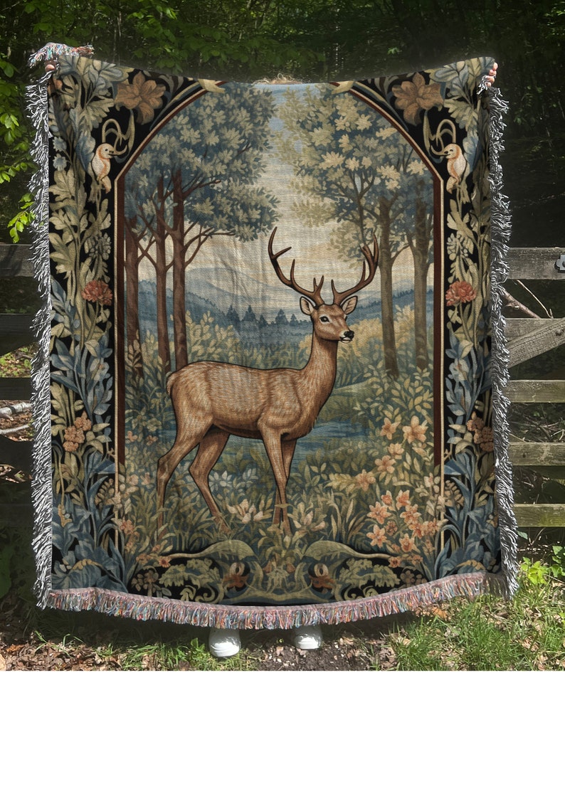 May include: A tapestry throw blanket with a vintage design featuring a deer standing in a forest. The deer is brown with large antlers and is surrounded by green trees and flowers. The tapestry is framed by a black border with intricate floral designs.
