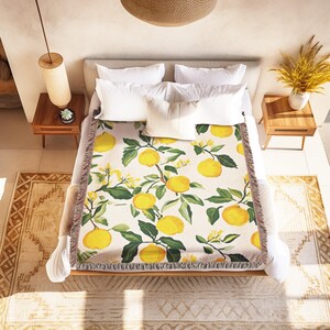 May include: A white throw blanket with a lemon print. The blanket has a fringe border and is on a bed with white pillows.