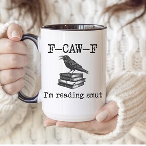 May include: White ceramic mug with a black interior and handle. The mug features a raven illustration perched atop a stack of books, with the text "F-CAW-F" above and "I'm reading smut" below.