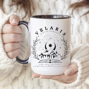 Può includere: Una tazza in ceramica bianca con bordo e manico neri. La tazza presenta un'illustrazione in bianco e nero di una catena montuosa, una luna crescente, una stella e un libro con il testo "VELARIS CITY OF STARLIGHT" e "To the stars who listen and the dreams that are answered".