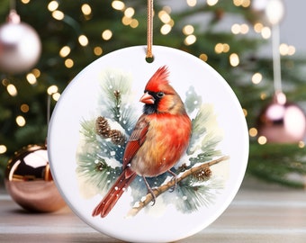 May include: A round, white Christmas ornament featuring a watercolor painting of a vibrant red cardinal perched on a snow-covered pine branch. The bird has a red crest and is set against a backdrop of evergreen boughs and pinecones. A gold string is attached for hanging.