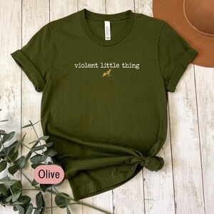 Puede incluir: Camiseta verde oliva con mangas enrolladas y dobladillo anudado. La camiseta presenta el texto "violent little thing" en una fuente serif, con un peque&ntilde;o gr&aacute;fico de perro. La palabra "Olive" est&aacute; en un &oacute;valo rosa.